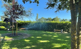 Grüner Park mit Rasenfläche, Bäumen, Holzbänken und einer kleinen Holzkonstruktion unter blauem Himmel.