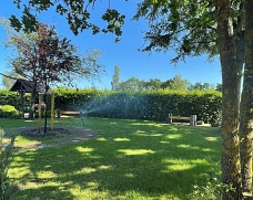 2026_Neues Urnenerdröhrenfeld_Weyhausen_1 Grüner Park mit Rasenfläche, Bäumen, Holzbänken und einer kleinen Holzkonstruktion unter blauem Himmel.