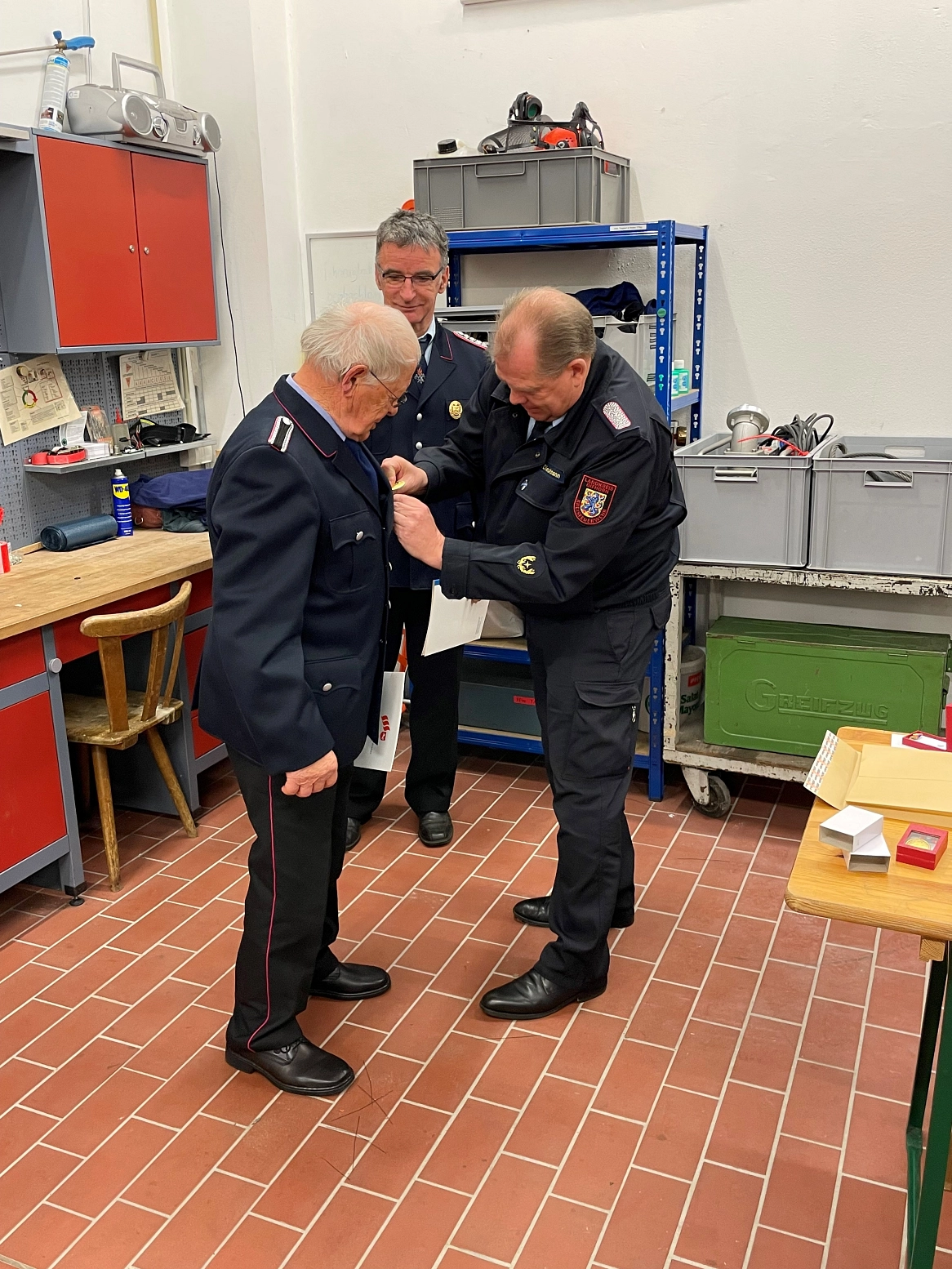 Der Kreisbrandmeister steckt dem Geehrten, einem weißhaarigen Mann in Uniform, eine goldene Ehrennadel an