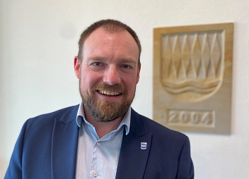 Mann Ende 30 mit gestutztem Vollbart, hoher Stirn und sehr kurzen braunen Haaren, lächelt offen in die Kamera, trägt blaues Sakko und ein hellblaues Hemd