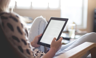 Person sitzt in einem Sessel und hält ein Tablet mit Textanzeige in den Händen.