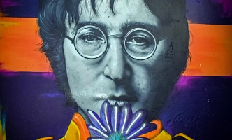 Porträt von John Lennon mit Brille vor farbigem Hintergrund, der eine Blume vor den Mund hält.