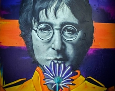 Wandbild von John Lennon Porträt von John Lennon mit Brille vor farbigem Hintergrund, der eine Blume vor den Mund hält.