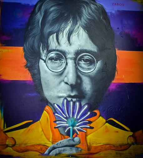 Porträt von John Lennon mit Brille vor farbigem Hintergrund, der eine Blume vor den Mund hält.